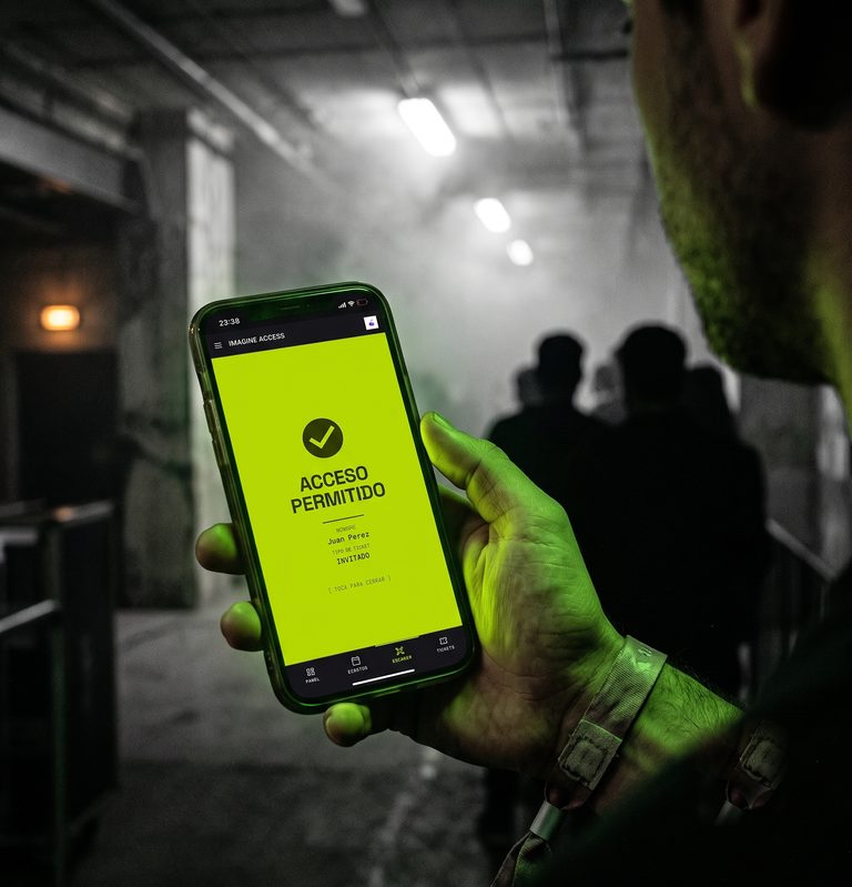 En la puerta de una fiesta en un galpón, una mano sostiene un teléfono que muestra ACCESO PERMITIDO en verde lima, con el nombre del invitado y su tipo de entrada